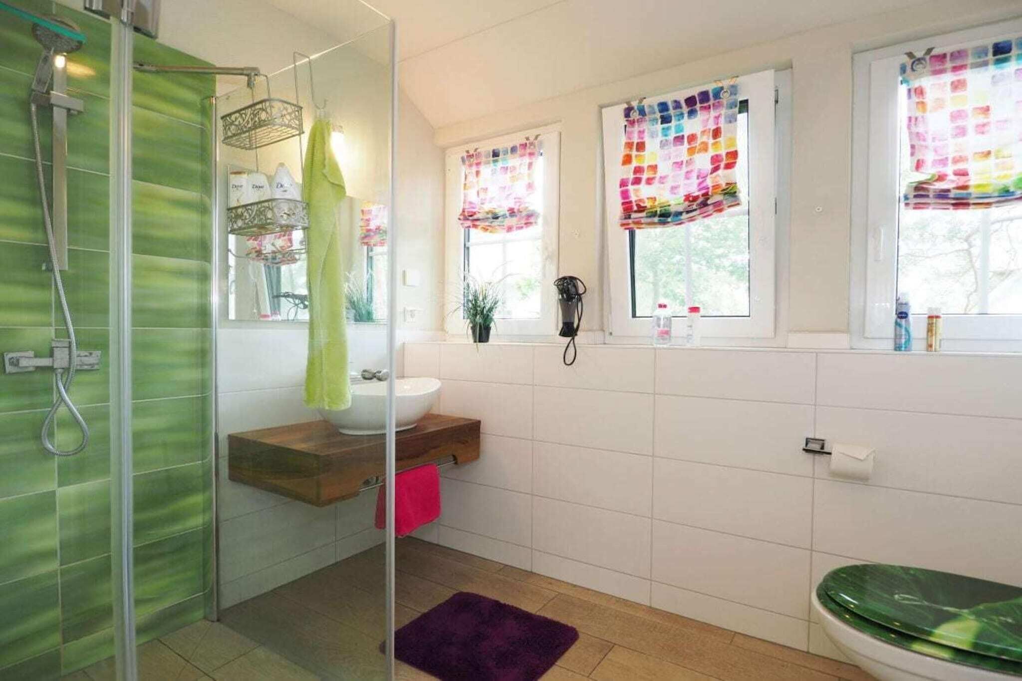 Ein Badezimmer mit Glasdusche, Holzwaschtisch, grüner Toilette und einem Fenster mit bunten Vorhängen.