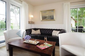A living room with a dark leather sofa, white armchairs, a wooden coffee table with a magazine, a candle, and a glass of champagne. - My Little Zanzibar 1 (Zirchow)