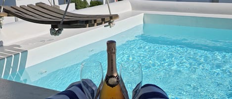 Una botella de champán Krone, dos copas y una toalla a rayas en un balcón con piscina y edificios blancos al fondo.