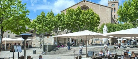 An outdoor cafe with tables and chairs, a stone building with a clock tower in the background, and a clear sky.