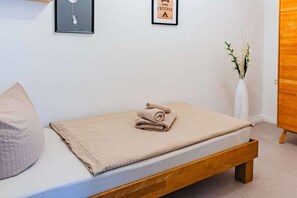 A single bed with a wooden frame, a beige blanket, and a rolled-up towel on top. There is a white vase with branches on the right side.
