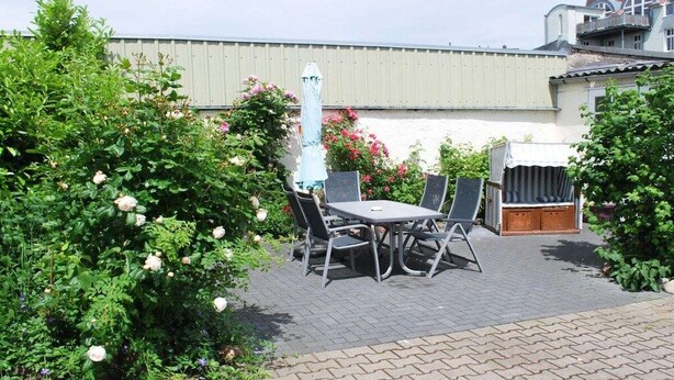 An outdoor patio area with a table and chairs, surrounded by lush greenery and flowering plants. - Holiday apartment with 2 bedrooms (Warnemünde)