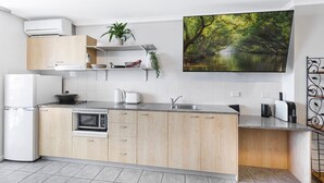 Une cuisine moderne équipée d’un four à micro-ondes encastré, d’une cuisinière et d’un évier. On y trouve un réfrigérateur blanc, une plante en pot et un tableau représentant une forêt accroché au mur.