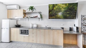 Eine moderne Küche mit integriertem Mikrowellenherd, einem Herd und einem Spülbecken. Ein weißer Kühlschrank, ein Blumentopf und ein Gemälde von einem Wald sind ebenfalls vorhanden.
