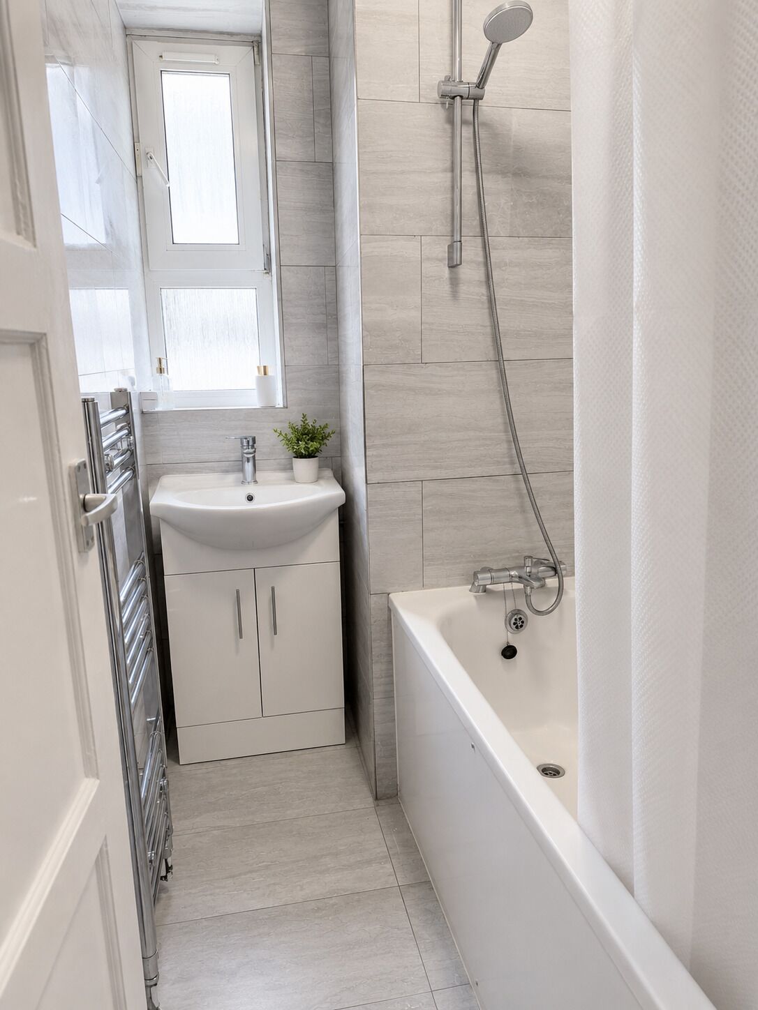 A narrow bathroom with a white sink, a mirror, a shower, and a door.