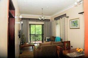Een woonkamer met een bank, een eettafel met fruit, een televisie en een balkon met gordijnen.