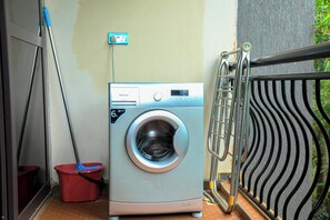 Eine kompakte Waschmaschine in einem kleinen Waschraum mit einem Metallregal und einem roten Eimer.