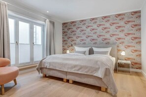 A bedroom with a bed, a nightstand, a lamp, a chair, and a window with curtains. - Bals Horsatal 15b / Rüm Hart (Wenningstedt)