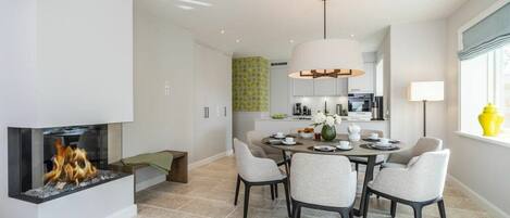 A modern dining area with a fireplace, a dining table with chairs, and a kitchen in the background.