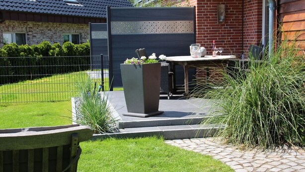 Eine Terrasse mit Tisch und Stühlen, einem Blumenbeet und einem Backsteinhaus im Hintergrund.
