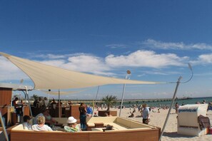 Eine Küstenlandschaft mit einer überdachten Sitzgelegenheit, einem Pier im Hintergrund und Menschen am Strand.