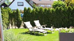 Ein Garten mit zwei Liegestühlen, einer Hecke und einem Gebäude im Hintergrund.