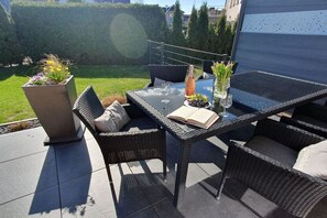 Een terras buiten met een tafel met glazen blad, rieten stoelen, een potplant en een fles wijn.