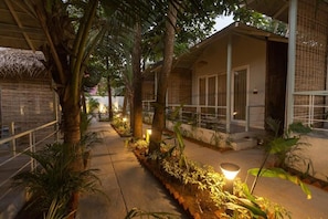 A pathway lined with trees and plants leading to a building with a thatched roof and a balcony. - Royal Touch Beach Huts (Canacona)
