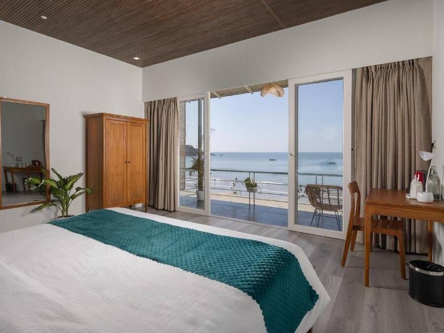 A bedroom with a bed, a wooden wardrobe, a desk with a chair, and a view of the sea through large sliding doors.