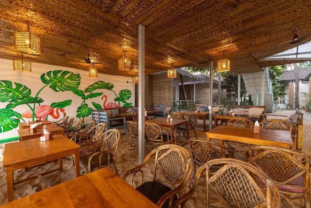Un espace de restauration intérieur avec des tables en bois et des chaises en osier, une fresque représentant des plantes tropicales et des flamants roses, et un plafond recouvert de chaume.