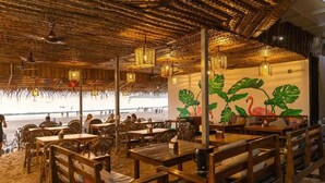 Un comedor interior con mesas y sillas de madera, vista al mar y un mural con plantas tropicales.