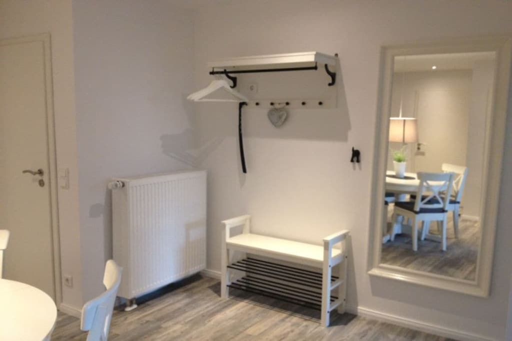 Une pièce avec un banc blanc, un radiateur, une chaise blanche et un miroir qui reflète un coin repas.