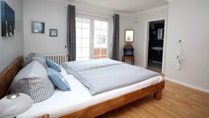 A bedroom with a large bed, a radiator, a window with curtains, a small table, and a door leading to a bathroom. - Holiday apartment in Sylt with 2 bedrooms (Sylt \/ OT Westerland)
