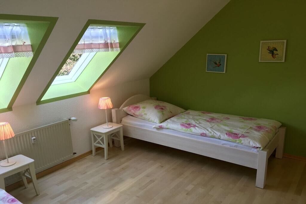 Une chambre avec un plafond en pente, deux fenêtres ornées de rideaux en dentelle, un lit avec une literie à motifs floraux, une table de chevet avec une lampe et un radiateur.