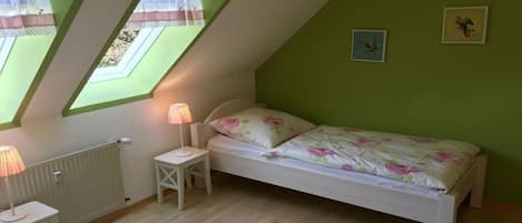 A bedroom with a sloped ceiling, two windows with lace curtains, a bed with floral bedding, a bedside table with a lamp, and a radiator.