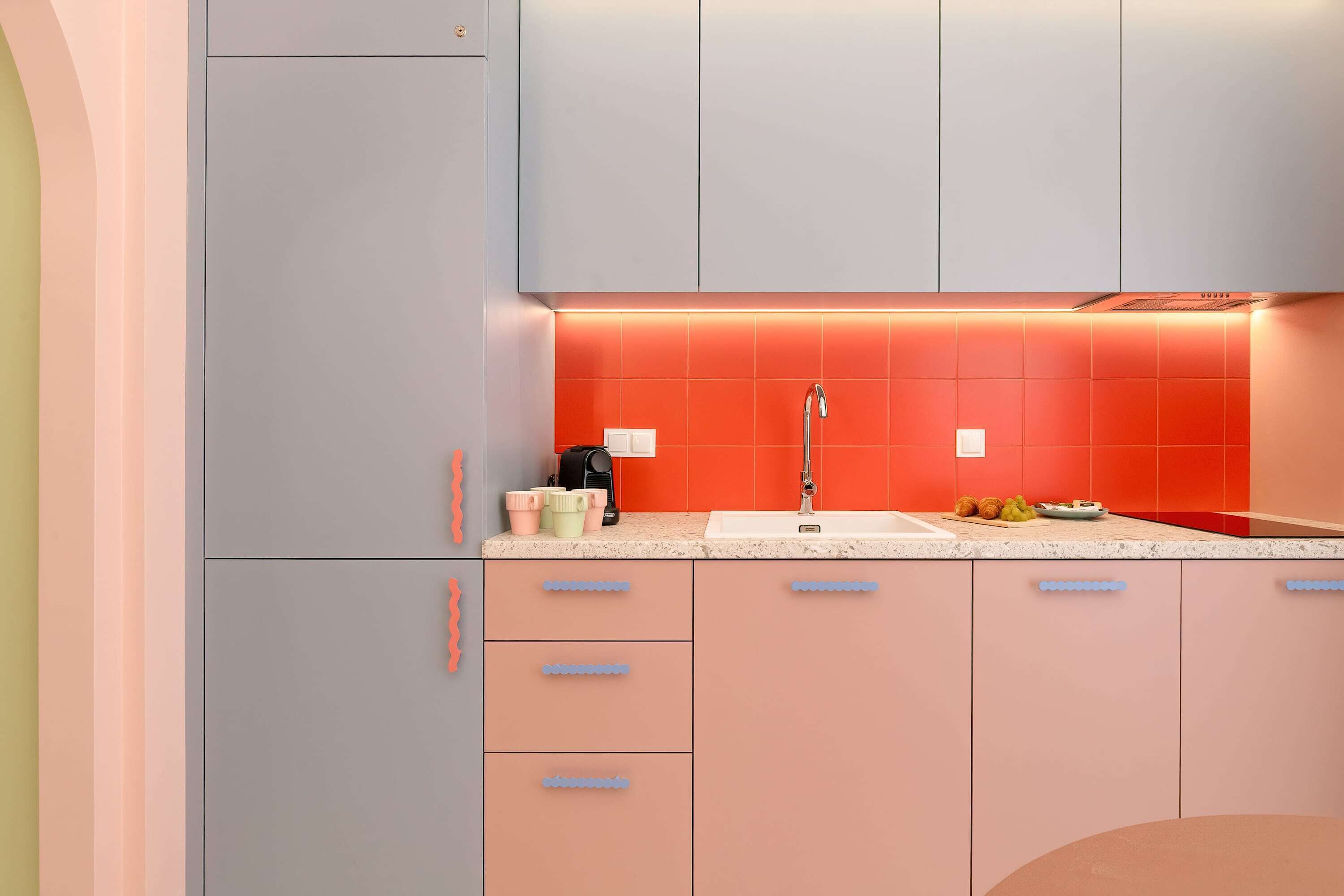 A modern kitchen with a grey refrigerator, a sink with a red backsplash, and a countertop with a coffee maker and cups.