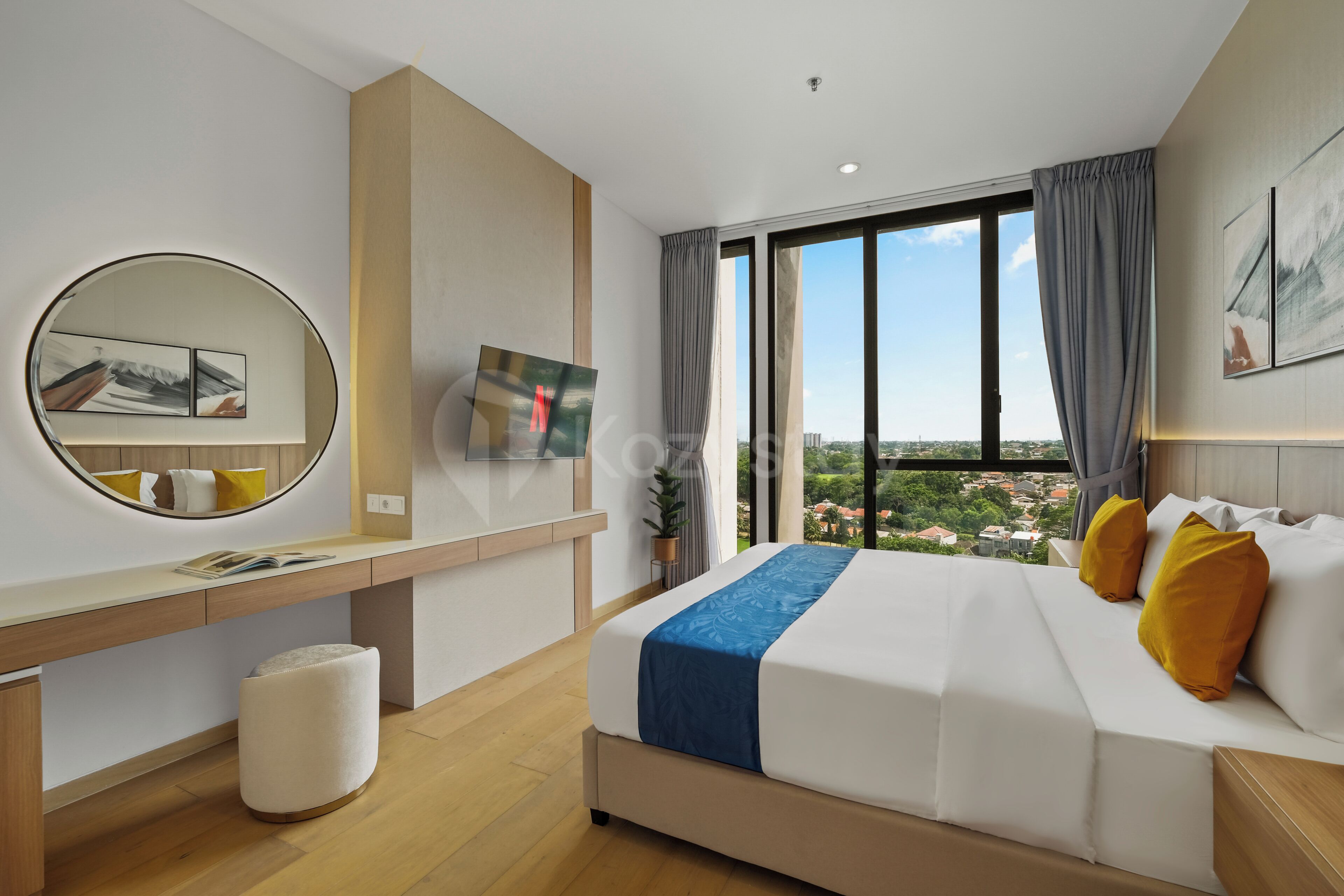 A modern hotel room with a large bed, a desk, a round mirror, and a view of the city through large windows.