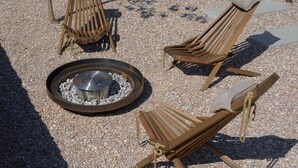 Drei hölzerne Lounge-Stühle mit Kissen, gruppiert um eine runde Feuerstelle mit Steinen.