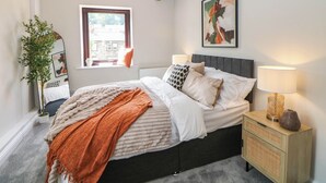 A bedroom with a bed, bedside lamps, a window, a plant, and a painting on the wall. - Half Under (Huddersfield)