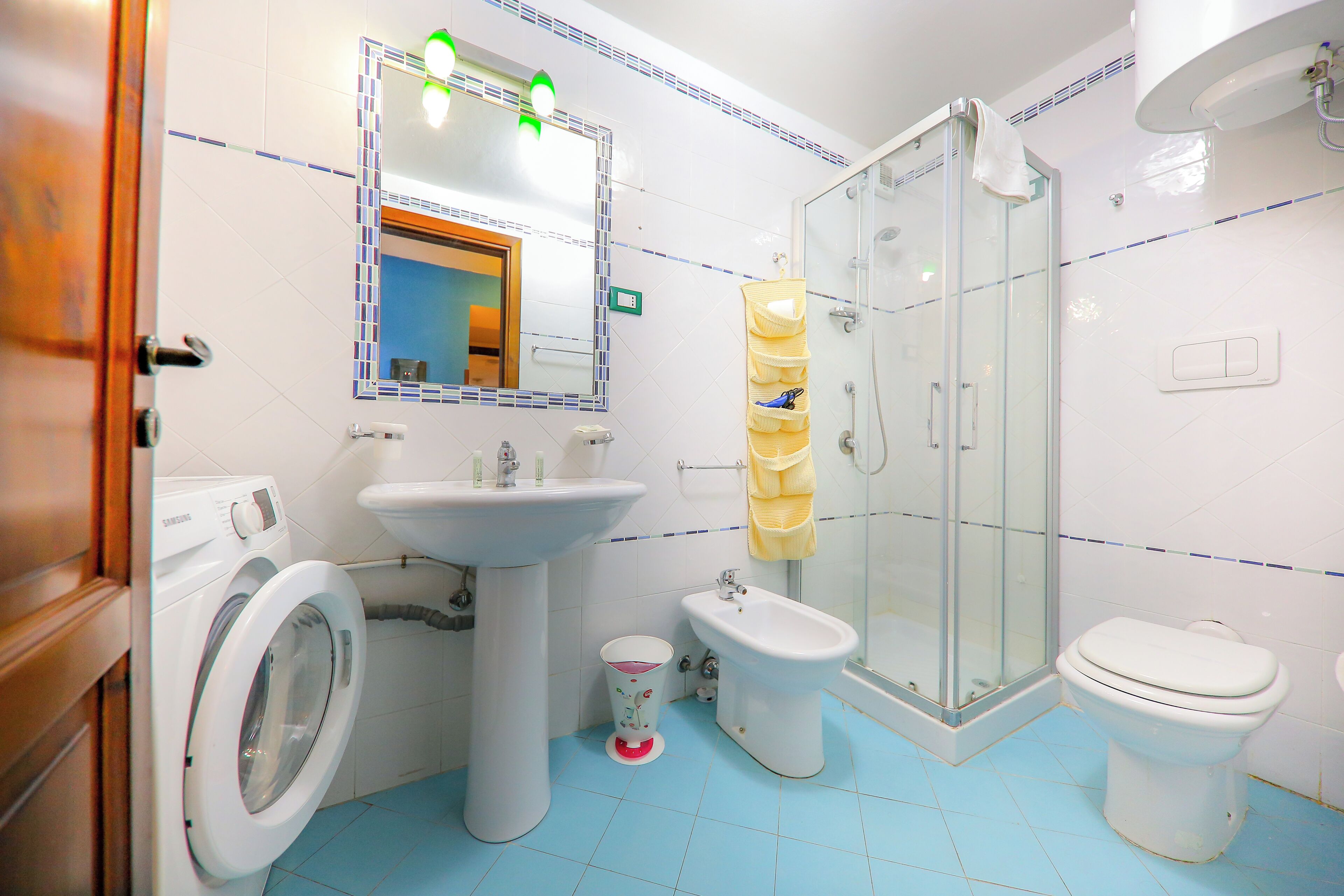 Et badeværelse med vaskemaskine, håndvask, toilet, bidet og bruseniche med glasvægge.