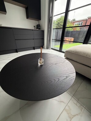 A modern living room with a round black coffee table, a beige sofa, and a view of a backyard through large windows.