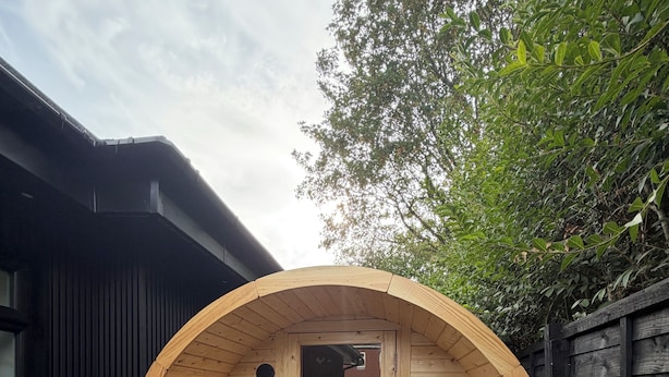 A wooden sauna structure with benches and a door, surrounded by a dark fence and trees.