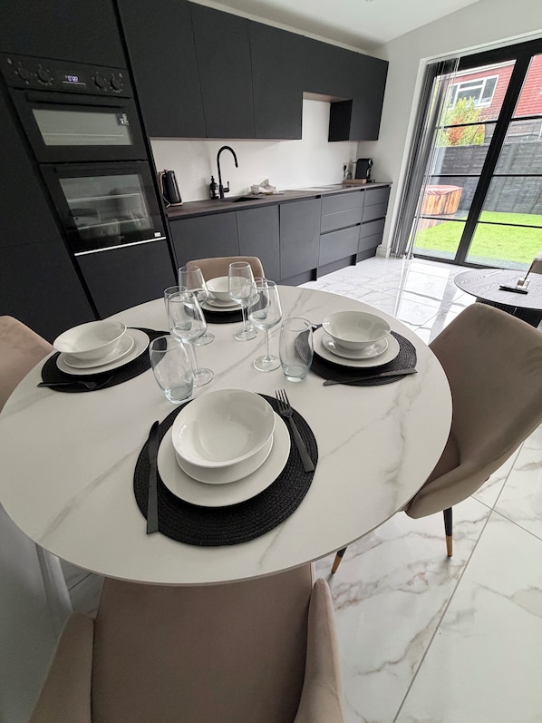 A modern kitchen with dark cabinetry, a dining table set with plates, glasses, and cutlery, and a view of a green lawn through a glass door.