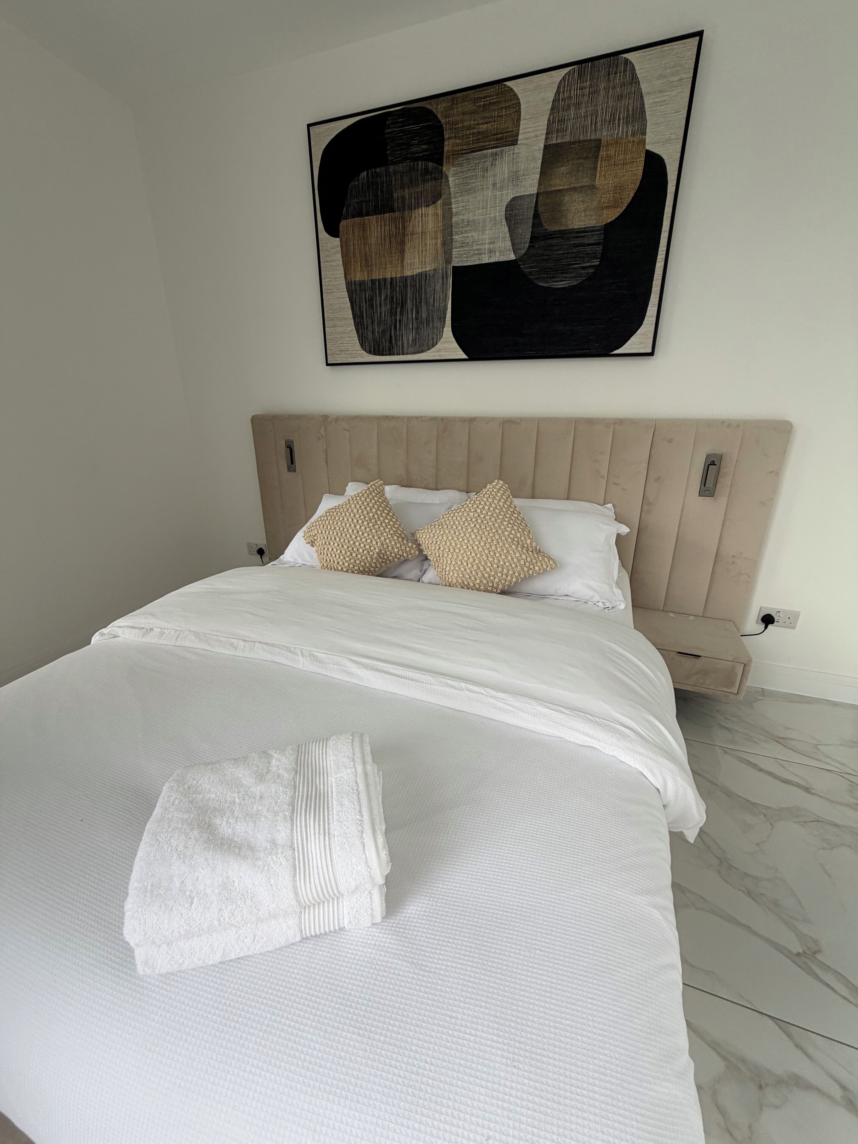A neatly made bed with a white comforter and two beige pillows, a wooden headboard, and a wall-mounted abstract painting.