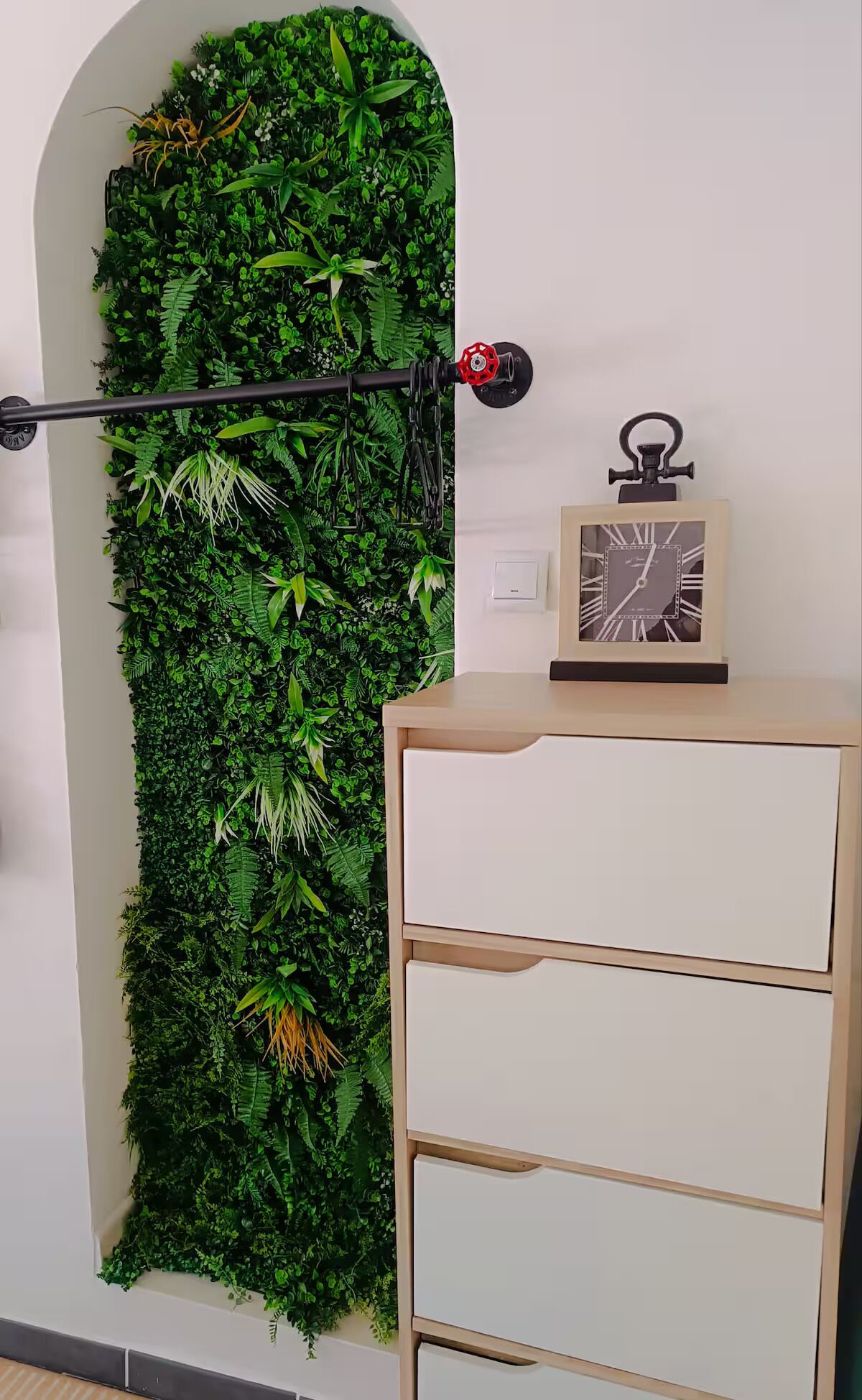A wall with a vertical garden, a black metal rod, and a clock on a cabinet.