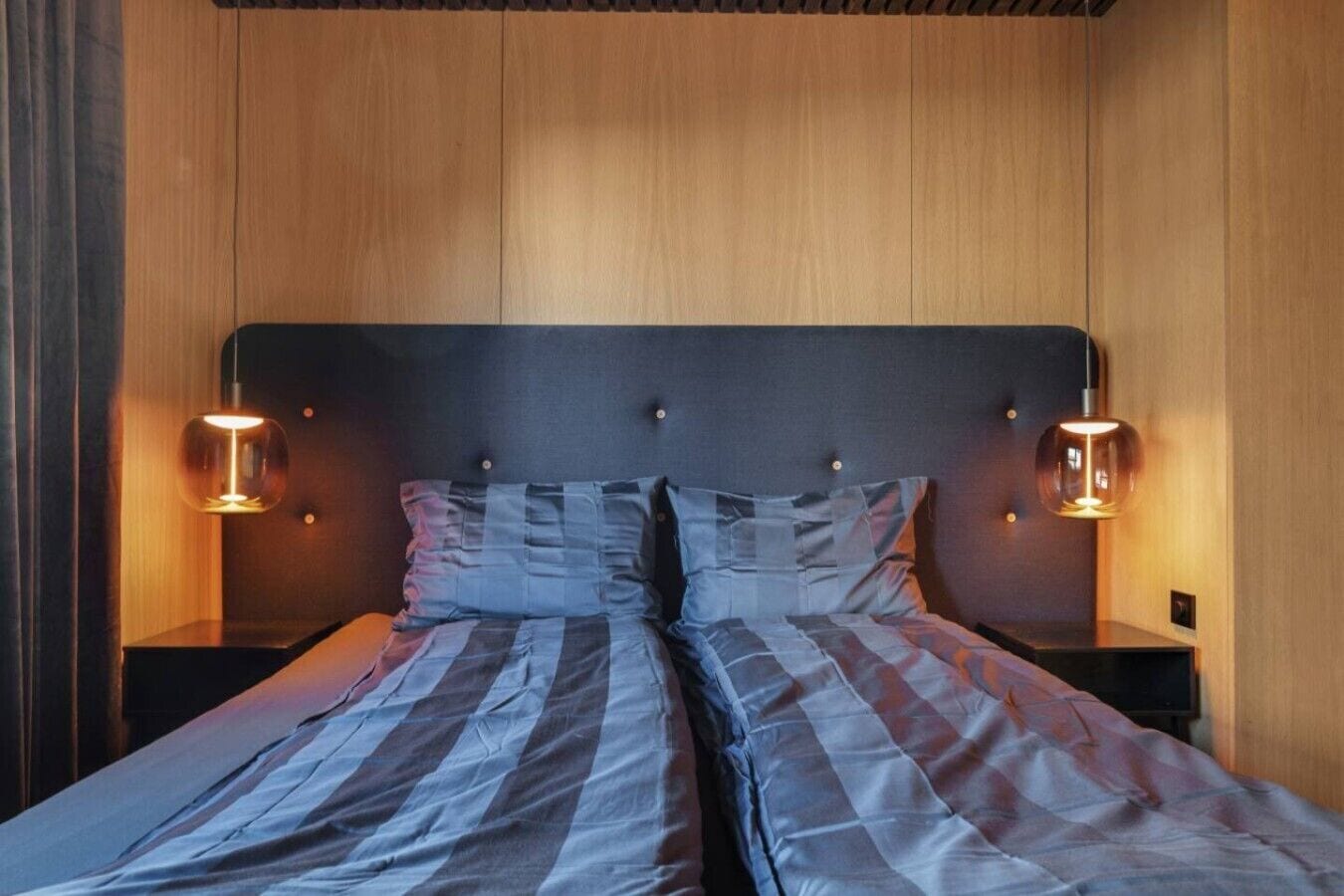 A double bed with a striped bedspread, two bedside tables with lamps, and wooden paneling on the walls.