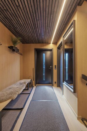 A hallway with a wooden ceiling, a bench, a rug, and a door with a vertical light feature. - Villa Panorama | Arctic Seasons | Luxury Living (Tromsø)