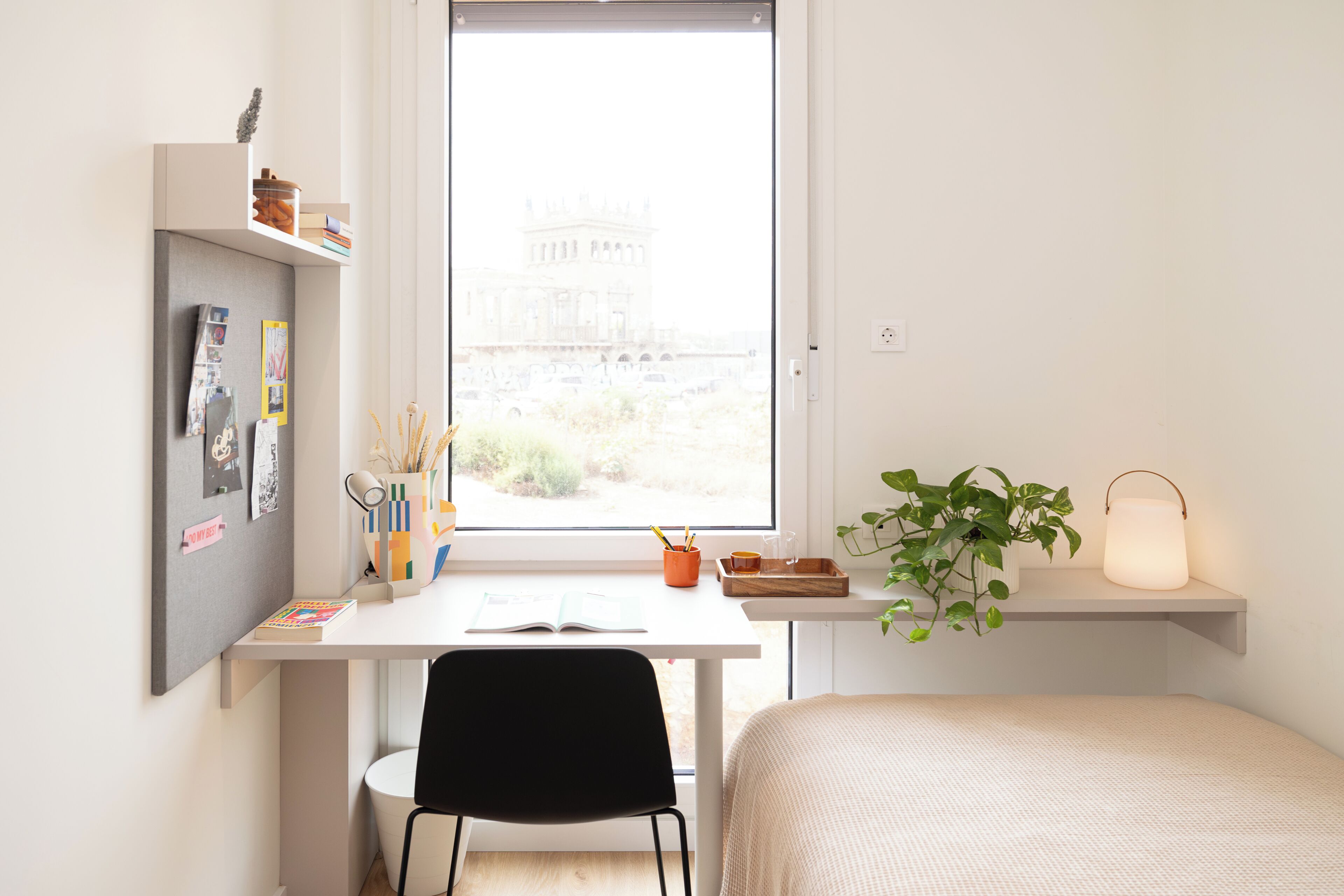 A minimalist bedroom with a bed, a desk with a chair, a window with a view of a building, and a shelf with a plant.