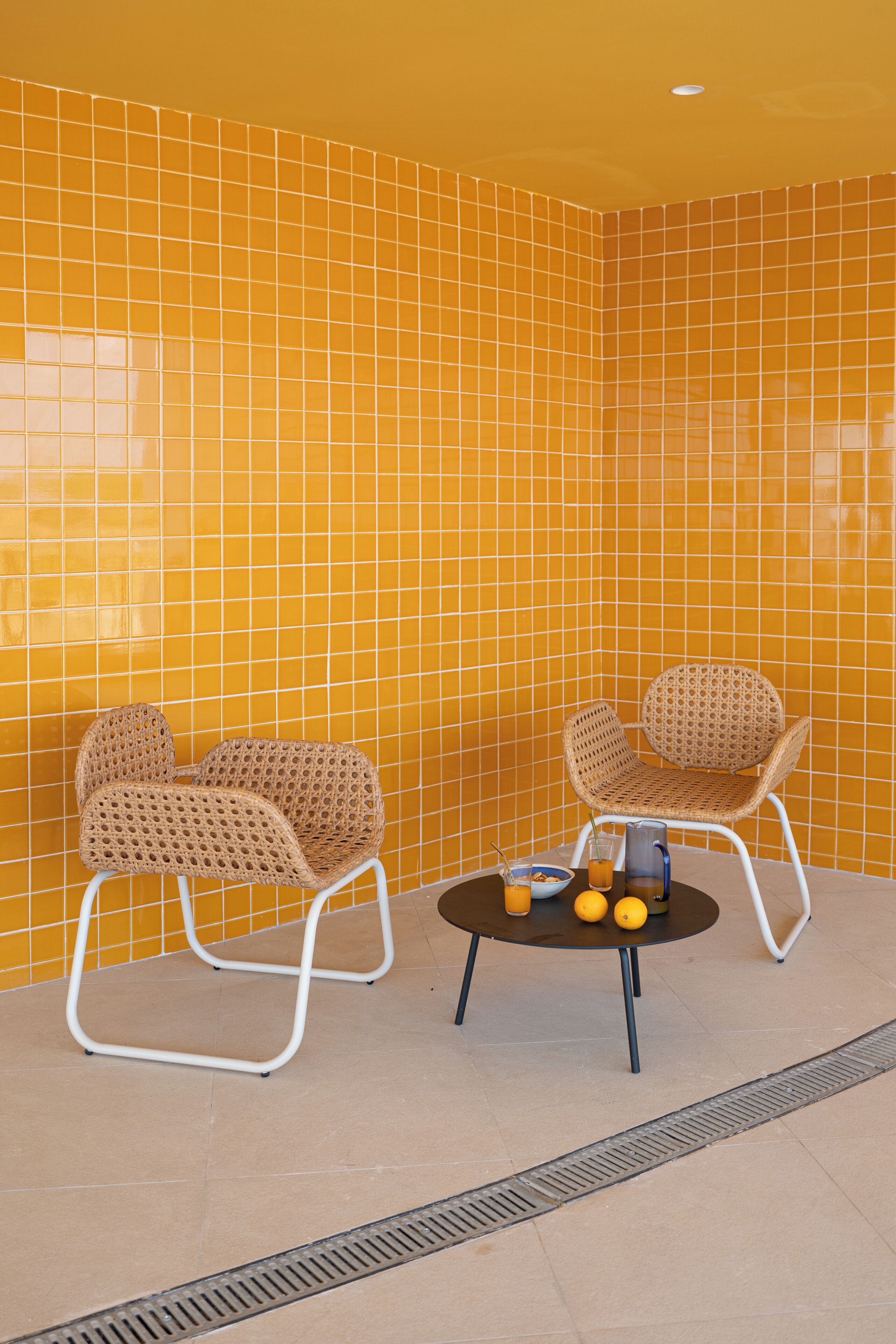A room with yellow tiled walls, two wicker chairs, a small round table with a bowl, two glasses, and two oranges.