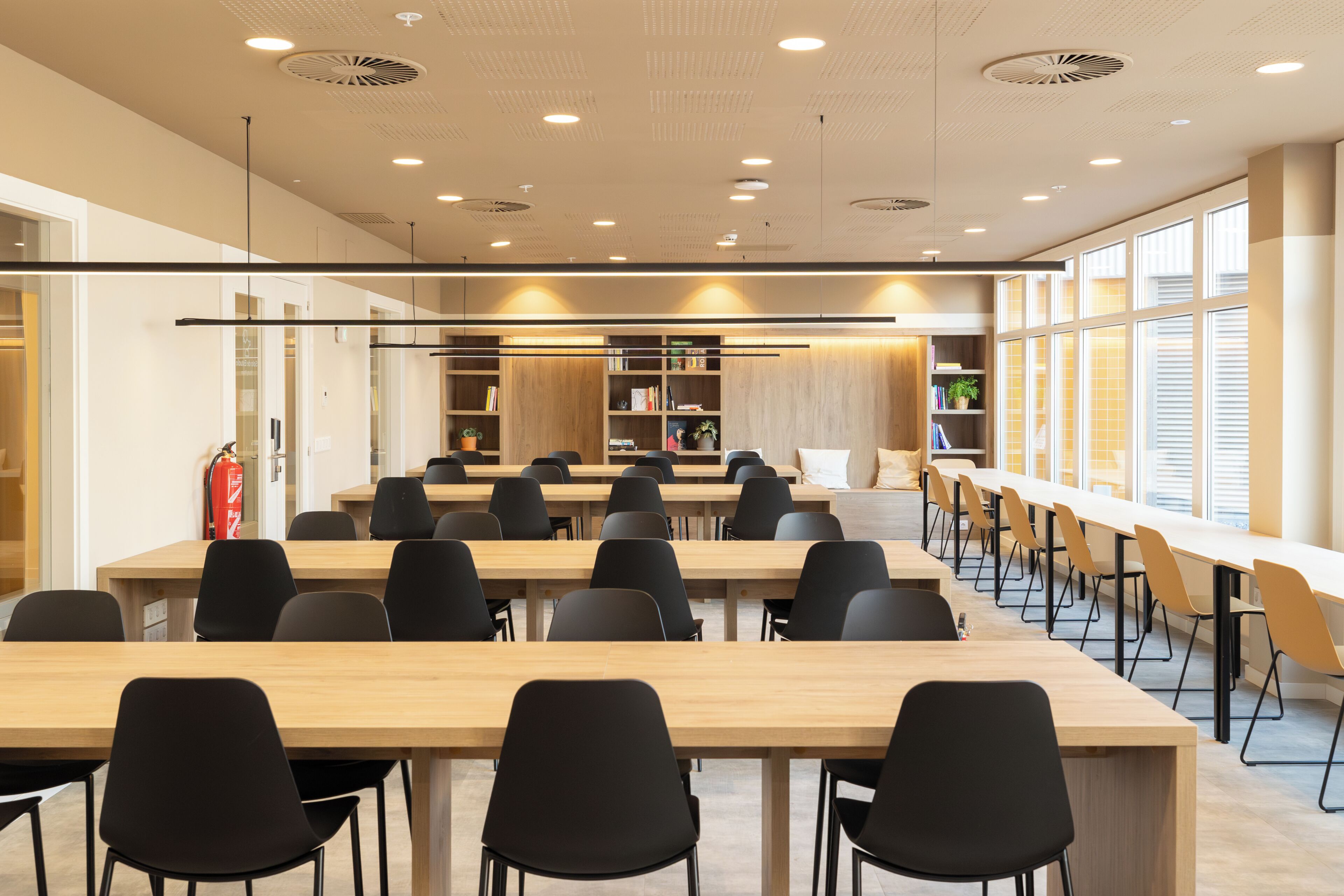 A spacious conference room with long tables and chairs, a wooden bookshelf, and a large window providing natural light.