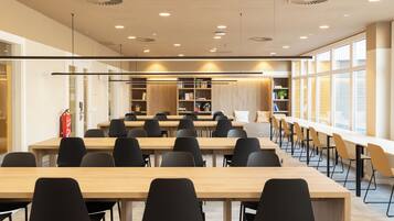 A spacious conference room with long tables and chairs, a wooden bookshelf, and a large window providing natural light.