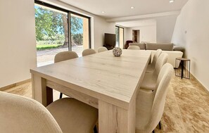 A modern dining area with a large wooden table, beige upholstered chairs, and a view of the outdoors through large windows. - Awesome home in St Martin de Crau (St Martin de Crau)