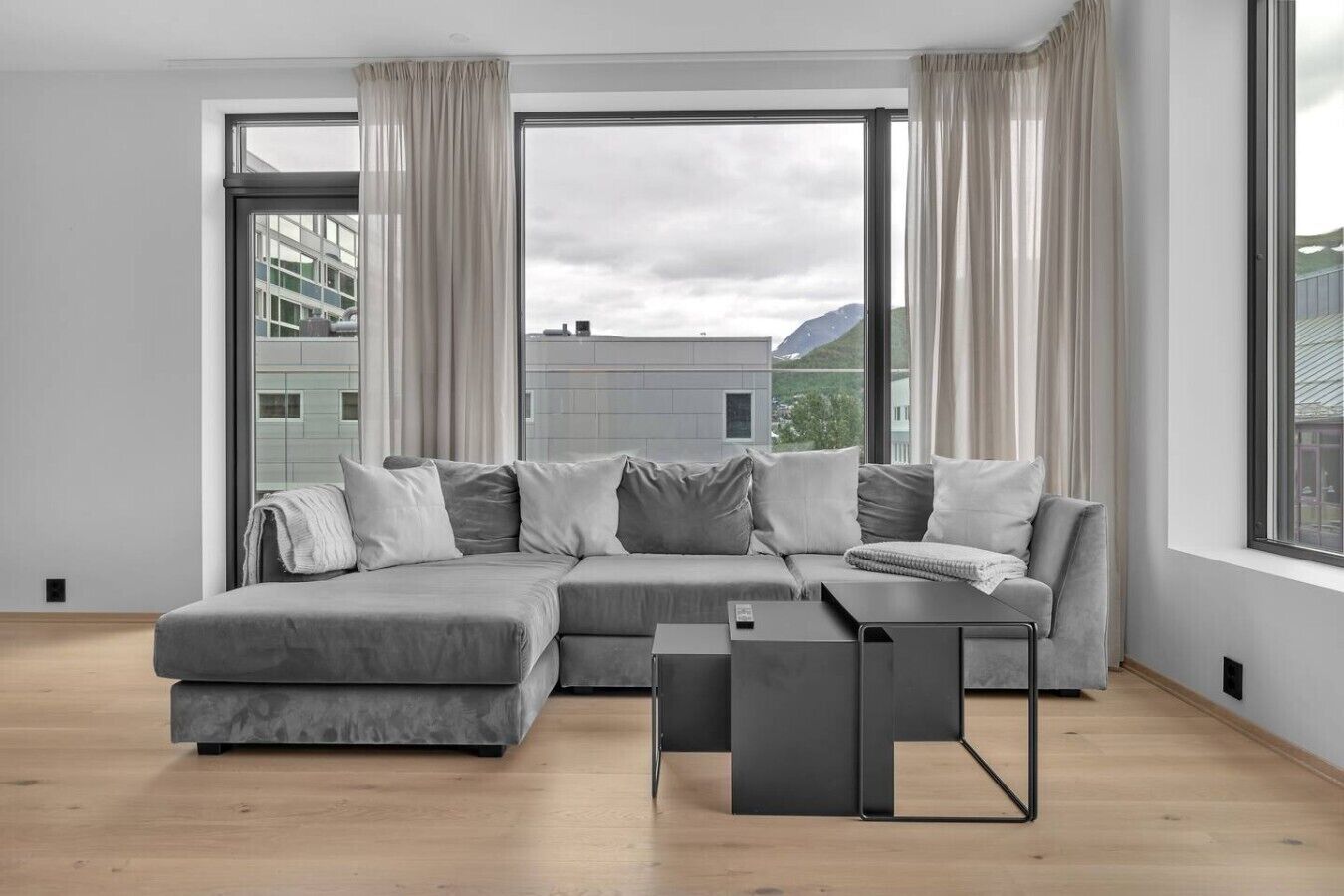 Un salon moderne avec un canapé d’angle gris, une petite table d’appoint et une vue sur les immeubles à travers de grandes fenêtres.