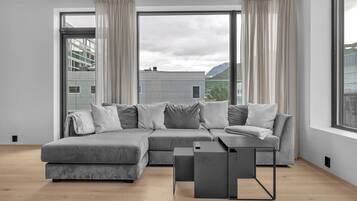 Un salon moderne avec un canapé d’angle gris, une petite table d’appoint et une vue sur les immeubles à travers de grandes fenêtres.