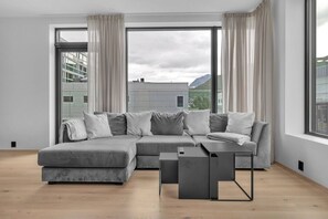 A modern living room with a grey sectional sofa, a small side table, and a view of buildings through large windows. - Arctic Urban Cityscape Sea & City Views, sleeps 4 (Tromsø)