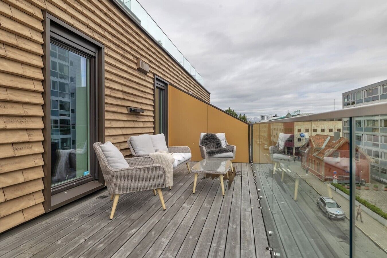 Une terrasse en bois avec des sièges extérieurs et une rambarde en verre donnant sur une rue.