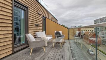 Une terrasse en bois avec des sièges extérieurs et une rambarde en verre donnant sur une rue.