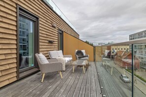 Um deck de madeira com área de estar externa e guarda-corpo de vidro com vista para a rua.