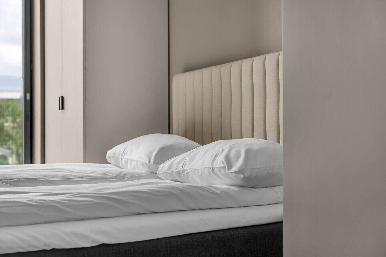 A bed with white bedding and a headboard with vertical lines, in a room with a view of greenery outside.