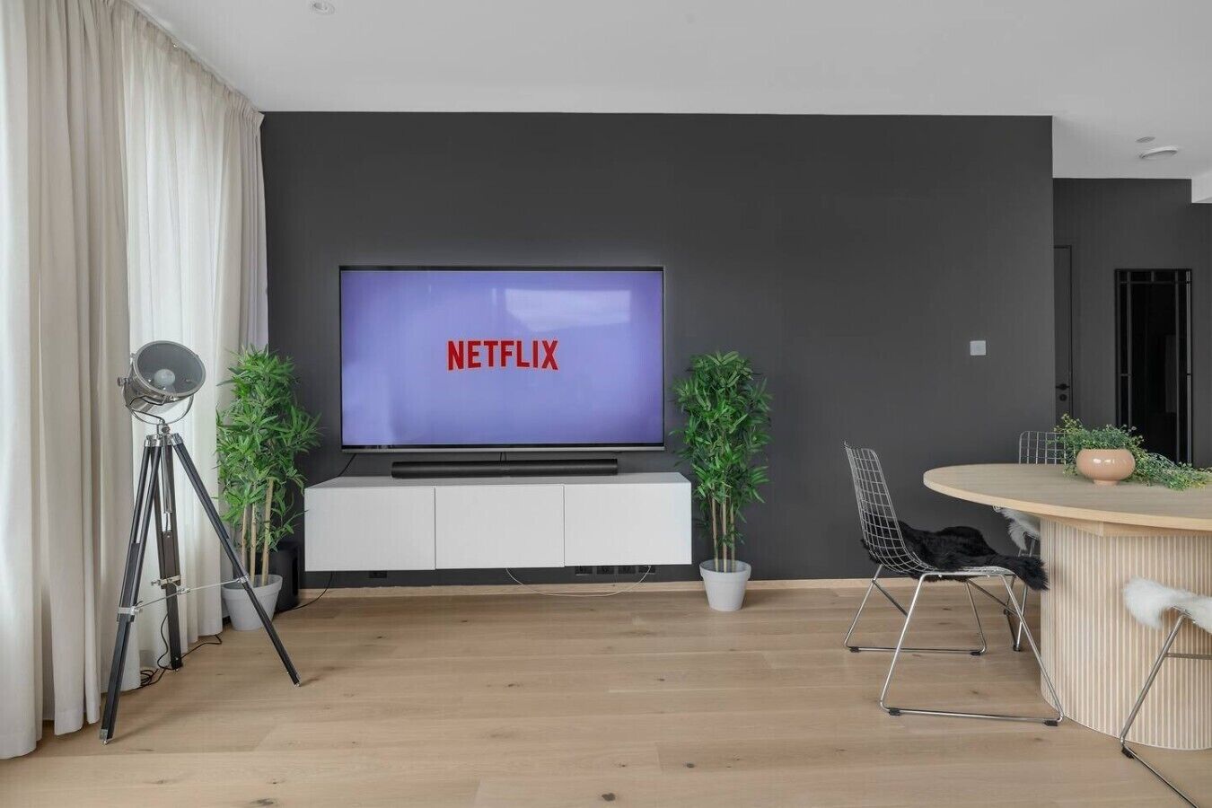 Ein modernes Wohnzimmer mit einem Flachbildfernseher, der 'NETFLIX' zeigt, einer weißen Unterhaltungseinheit, einem Stativ, zwei Topfpflanzen, einem runden Esstisch mit Stühlen und einer dunklen Wand.